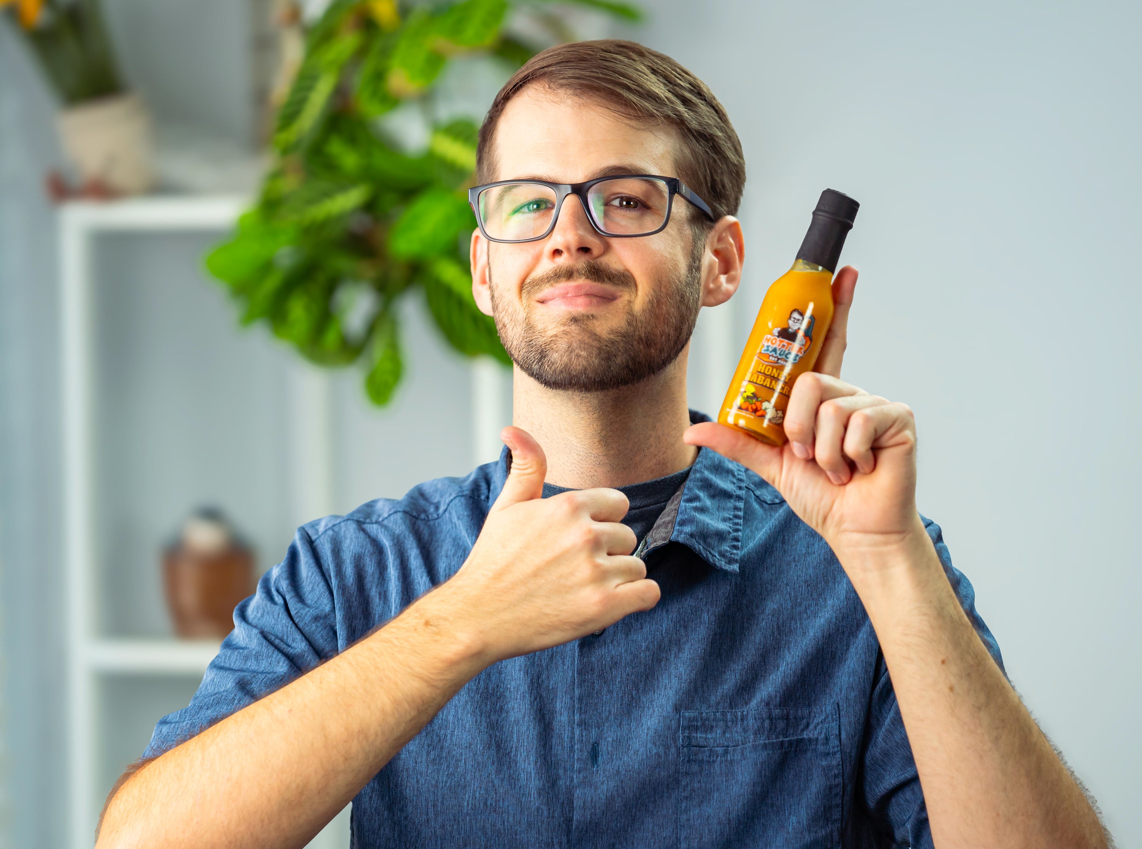 Man holding a bottle of hot sauce and giving a thumbs-up gesture indoors.