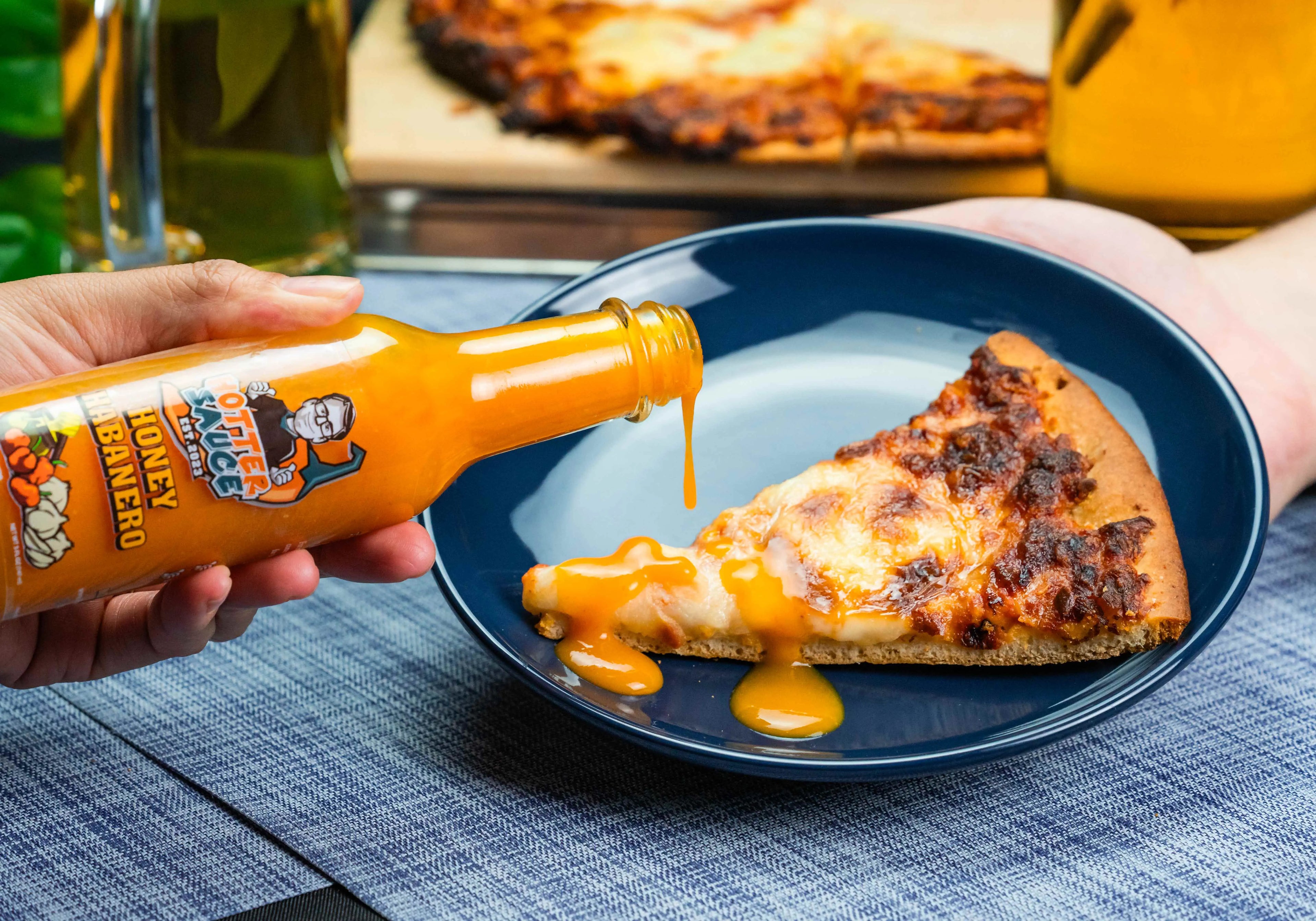 Person pouring honey habanero hot sauce onto a slice of pizza on a blue plate with a bottle of hot sauce in the foreground.