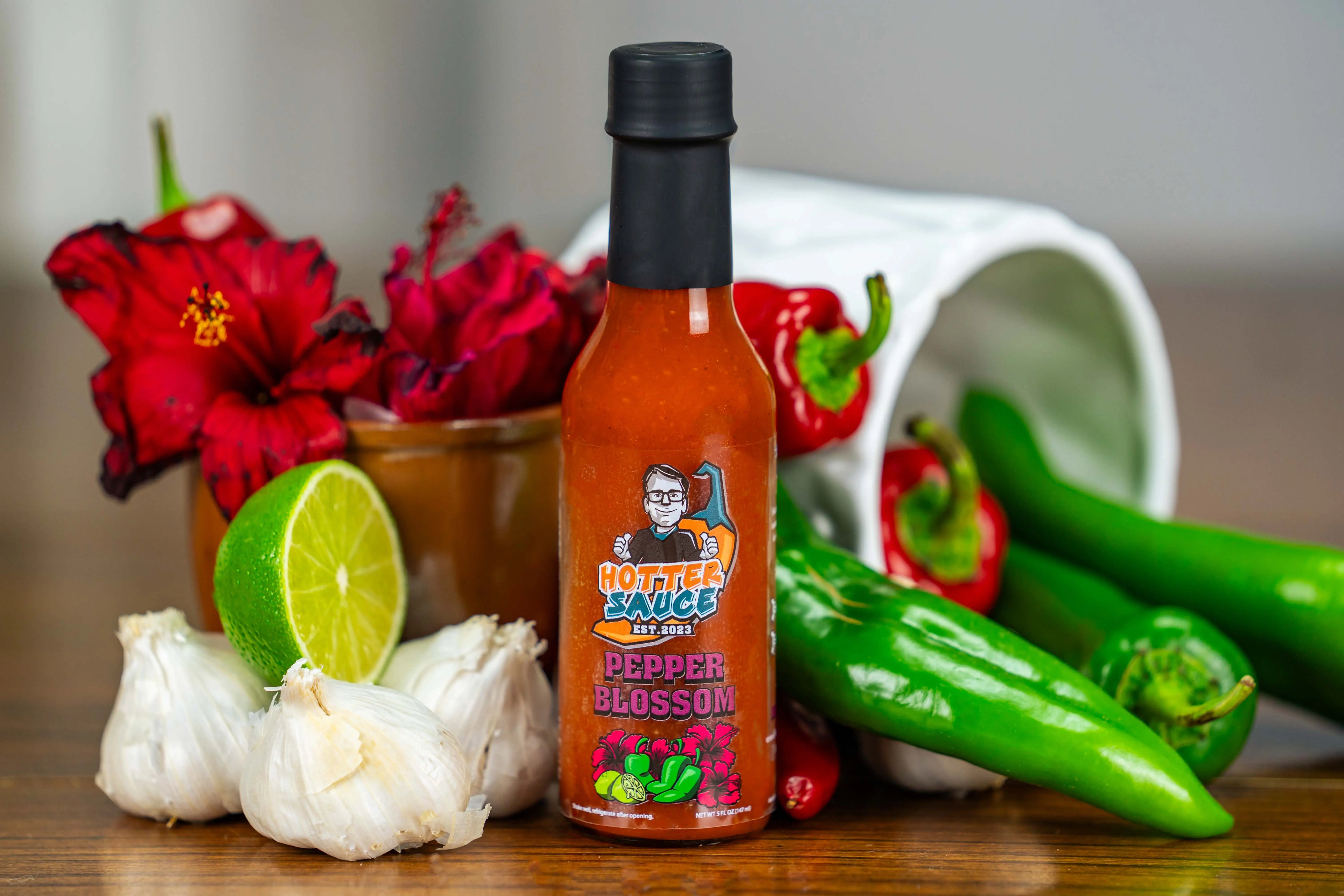 Pepper blossom hot sauce bottle with peppers, garlic, and flowers on a wooden surface
