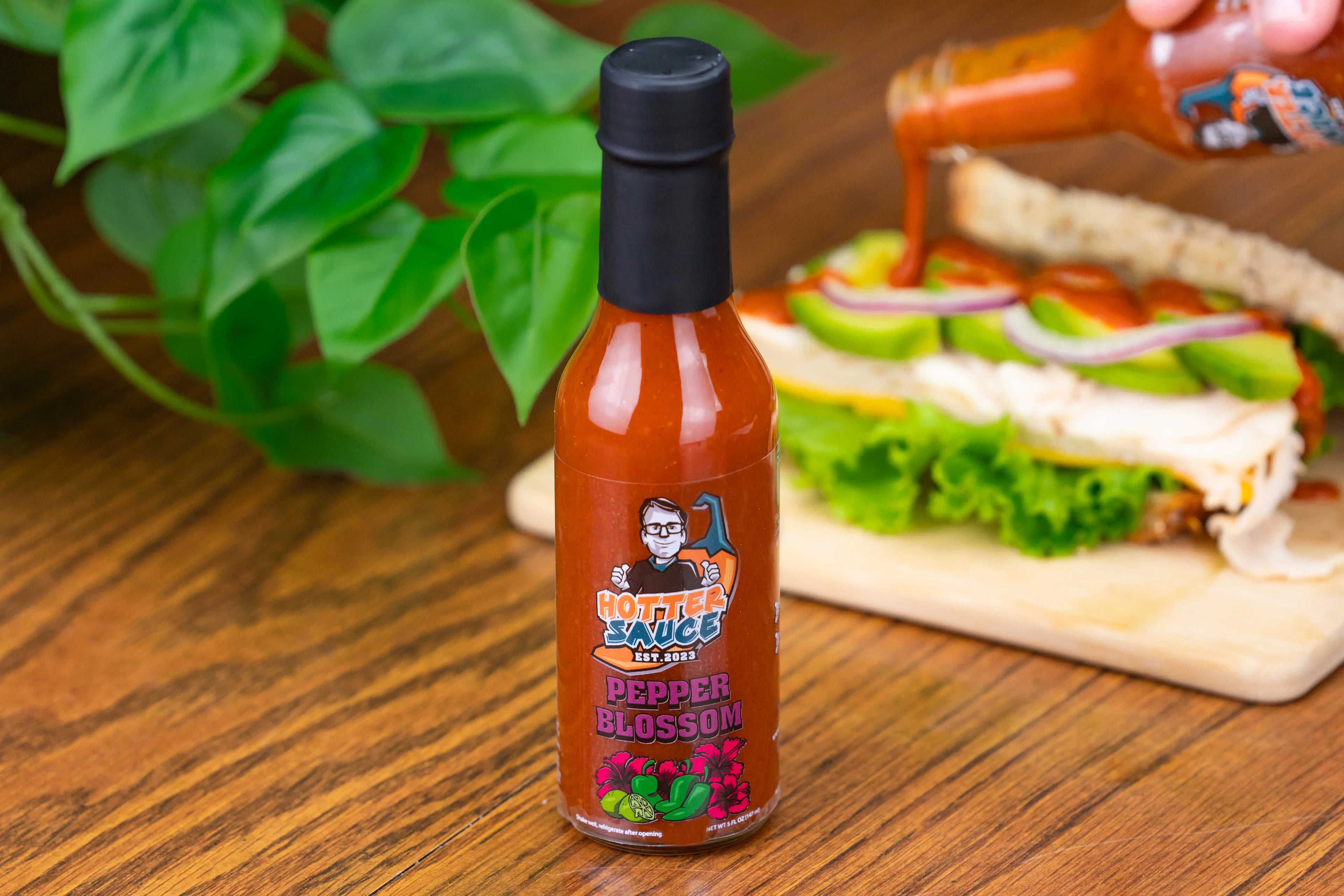 Hot sauce bottle labeled 'Pepper Blossom' on a wooden table with a sandwich in the background.