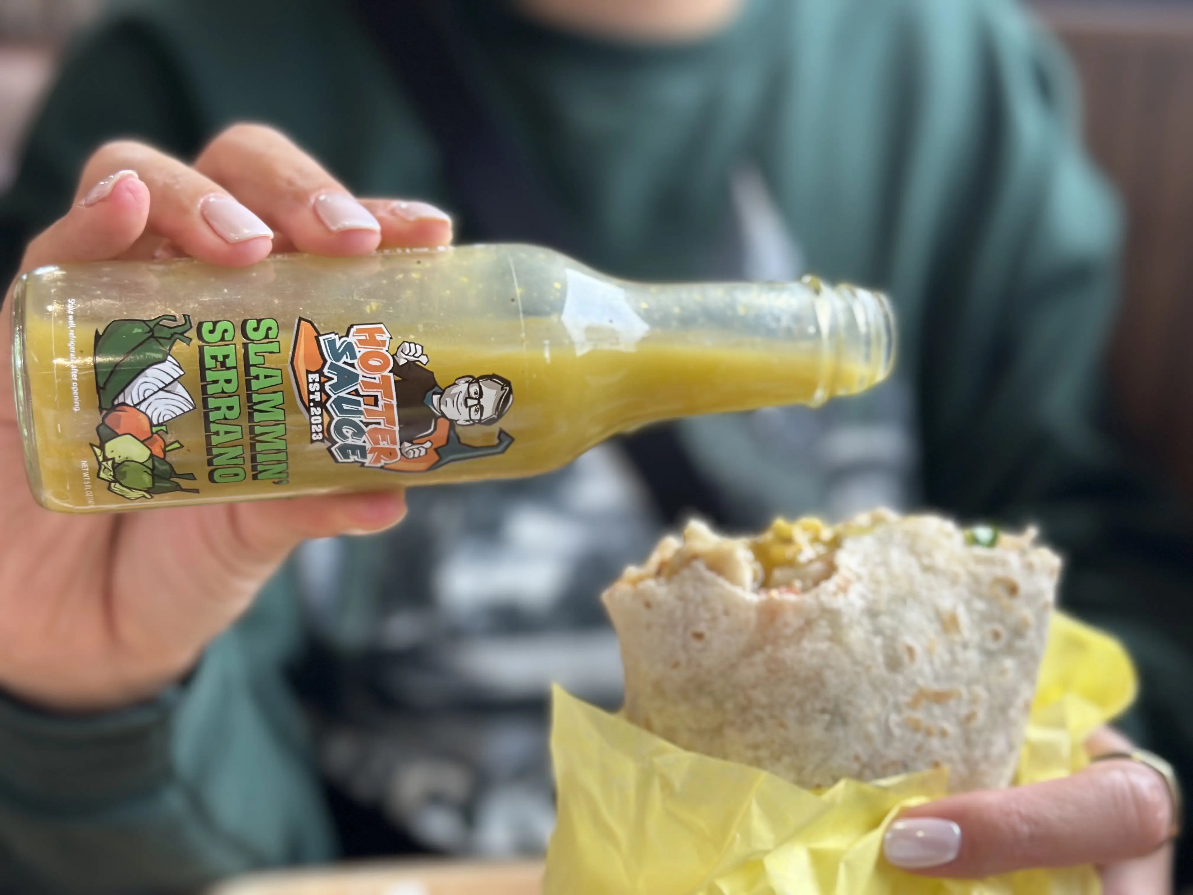 Hand holding a bottle of Slammin Serrano hot sauce with a burrito wrapped in yellow paper.
