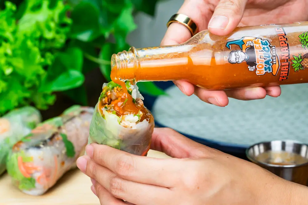 Person pouring Thai apple hot sauce from a bottle onto a spring roll with a green leafy background